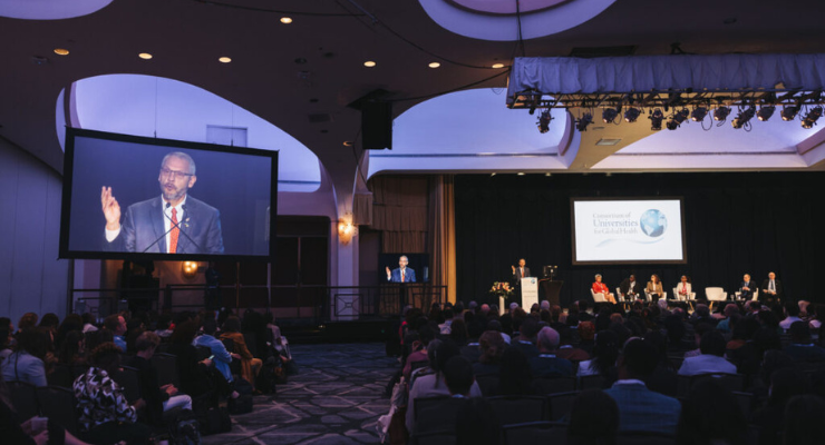 A man gives a speech in a large, dark ballroom with his image projected onto a screen