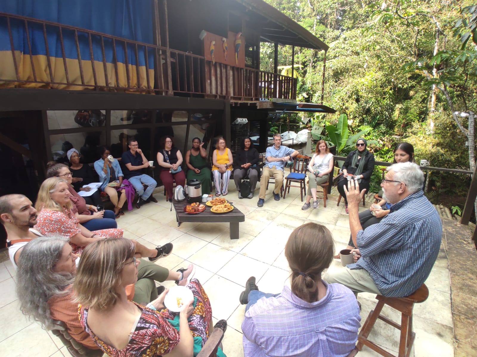 A group of people sit in a circle outside listening to a speaker