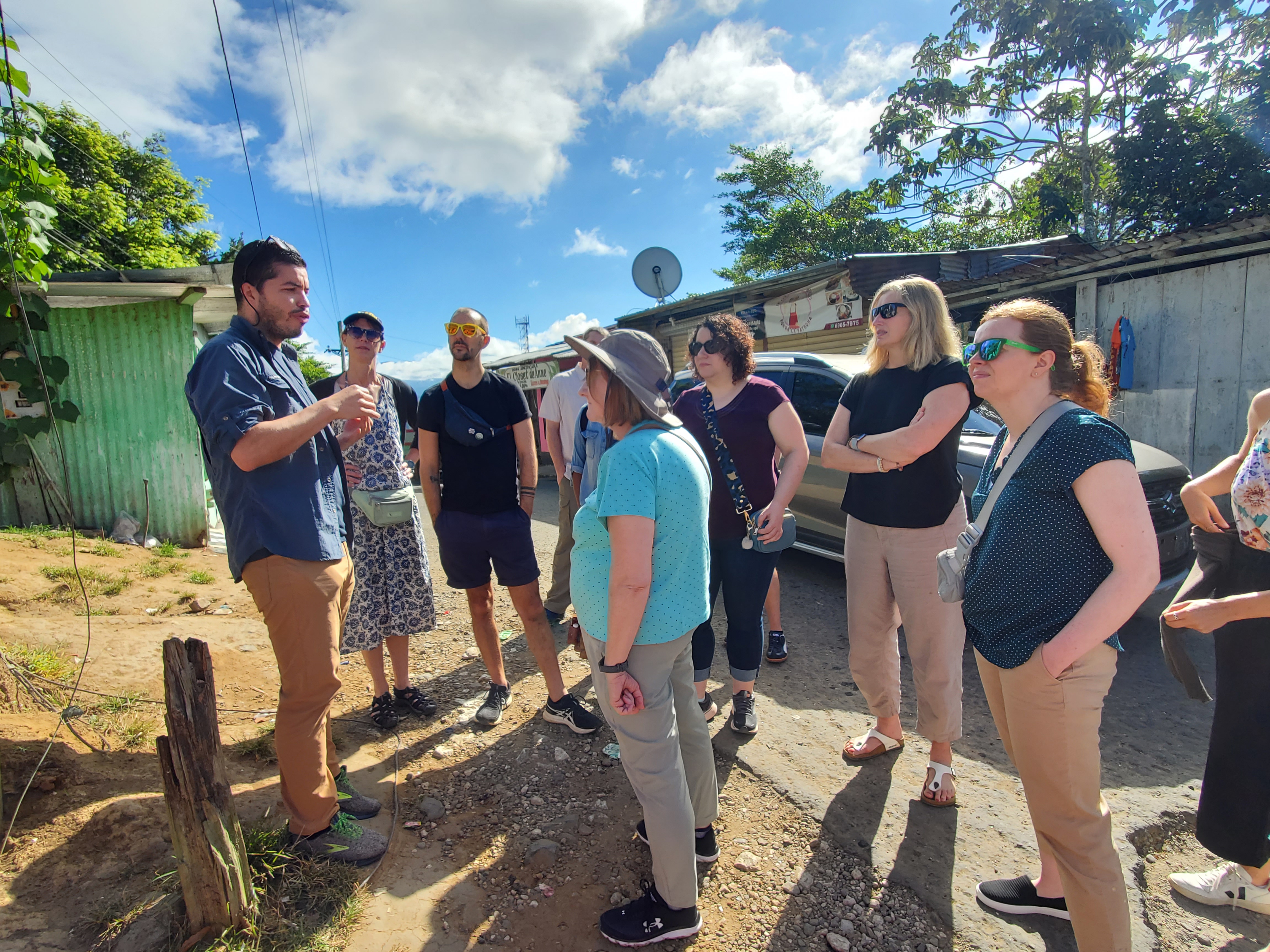 A group of people stand outside listening/watching a speaker