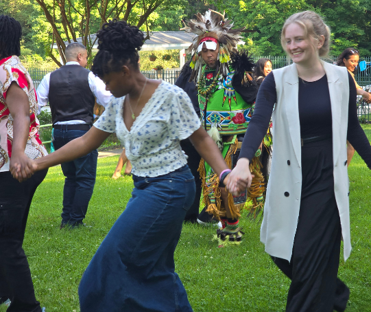 People dance in a circle around a man in Native American dress.