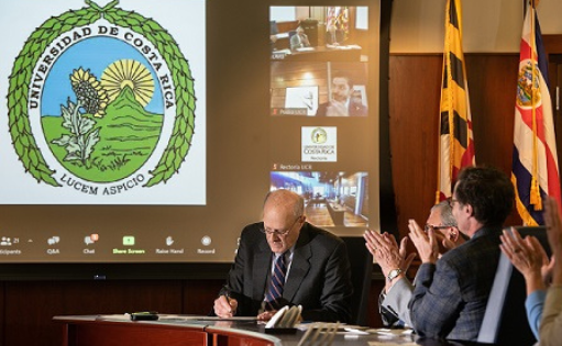 Dr. Bruce Jarrell signs an MOU in front of a screen with video from Costa Rica