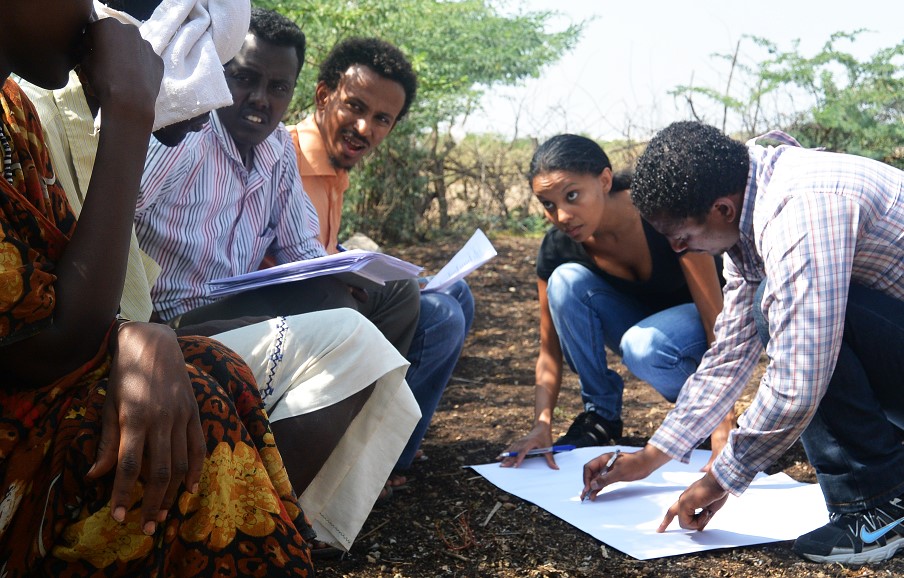 A man and woman squat to look at a large paper while others look on