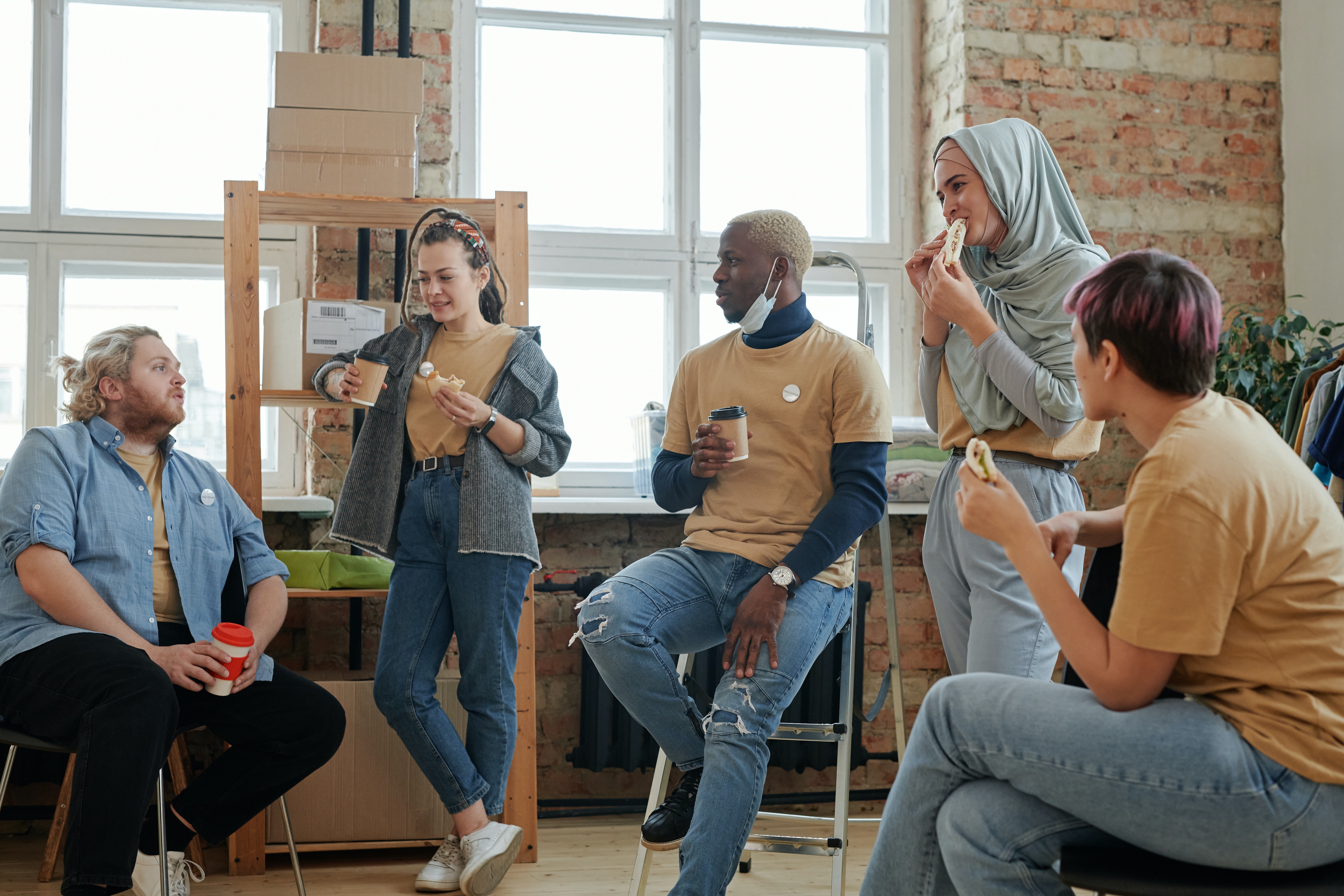 Young adults sit and stand in a group chatting and eating sandwiches.