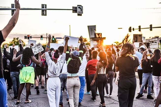 Large group of people marching away from the camera, some holding signs in protest